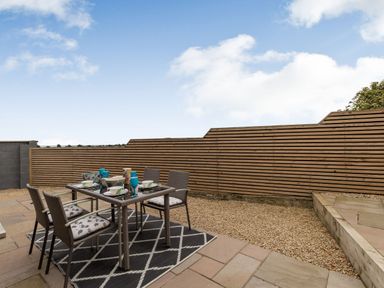 An outdoor patio with a glass table set with dishes and blue glasses on a rug and surrounded by four chairs by a wooden slat fence at Secret Kingdom Cottage in Warkworth