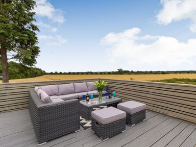 An outdoor deck with wicker furniture including a corner sofa two ottomans and a glass top table with glasses and flowers overlooking fields at Secret Kingdom Cottage in Warkworth