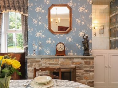 A dining area with a table set with plates and cutlery a stone fireplace with a clock and statue on the mantel and a window with curtains at Silver Crooks in Trewyn Nr Holsworthy
