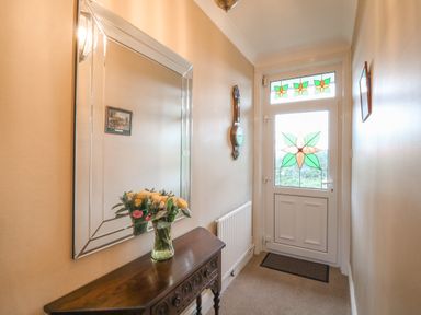 A hallway with a wooden table holding a vase of flowers a large mirror on the wall a clock a radiator and a door with stained glass at Silver Crooks in Trewyn Nr Holsworthy