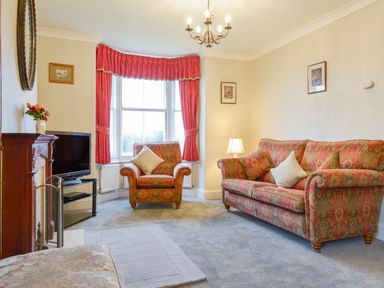 A living room with red patterned sofa and armchair near windows with red curtains at Silver Crooks in Trewyn Nr Holsworthy