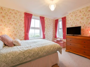 A bedroom with a double bed pink curtains floral wallpaper a wooden dresser and a television at Silver Crooks in Trewyn Nr Holsworthy