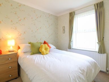 A bedroom with a bed white bedding colorful cushions wooden bedside table lamp and a window with curtains at Silver Crooks in Trewyn Nr Holsworthy