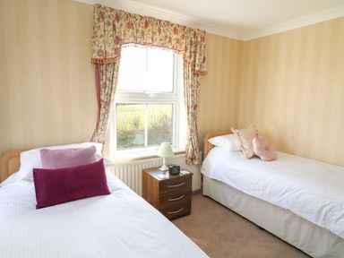 A bedroom with two single beds a wooden nightstand with a lamp and clock between the beds and a window with floral curtains at Silver Crooks in Trewyn Nr Holsworthy