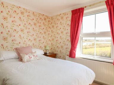 A bedroom with a bed white bedspread pink and butterfly cushions floral wallpaper a bedside table with a lamp and a window with red curtains at Silver Crooks Trewyn Nr Holsworthy