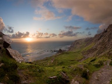 A coastal cliffside with grassy hills and a rocky path overlooking the ocean at sunset at Silver Crooks in Trewyn Nr Holsworthy