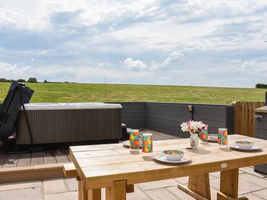 An outdoor patio with a wooden table set with plates glasses and a vase of flowers next to a hot tub and a grassy field at Silver Crooks in Trewyn Nr Holsworthy