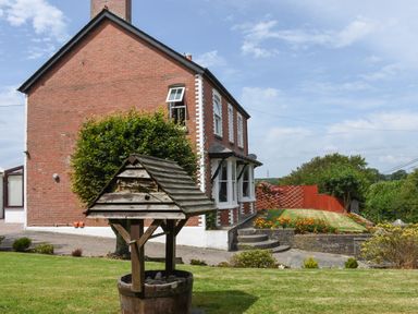 A brick house with white window frames and a wooden wishing well in the garden at Silver Crooks in Trewyn Nr Holsworthy