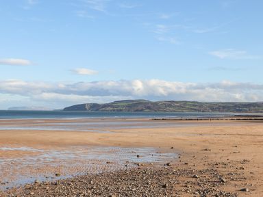 A sandy beach with scattered rocks and shallow water with hills and fields in the background at 4 Parc Delfryn in Brynteg near Benllech