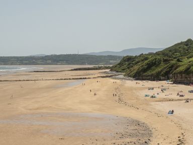 A sandy beach with people walking and sitting near a grassy hillside at 4 Parc Delfryn in Brynteg near Benllech
