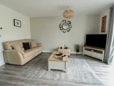 A living room with a beige sofa a coffee table a TV on a stand and a round wall mirror at 4 Parc Delfryn in Brynteg near Benllech