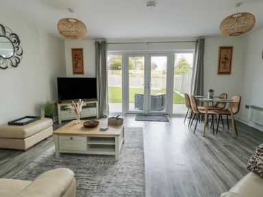 A living room with a television on a stand a coffee table a dining table with four chairs and glass doors leading to a garden at 4 Parc Delfryn Brynteg near Benllech