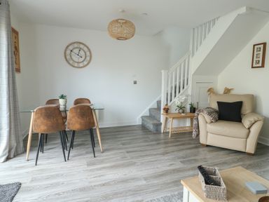 A dining area with a glass table and four chairs next to a sitting area with a beige armchair and a small wooden table under stairs at 4 Parc Delfryn Brynteg near Benllech