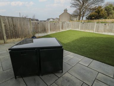 A backyard with a glass top table and two chairs on a stone patio and artificial grass at 4 Parc Delfryn in Brynteg near Benllech
