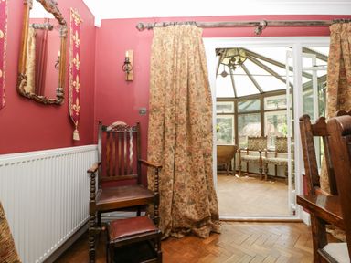 A dining area with wooden chairs and patterned curtains leading to a sunroom with upholstered chairs at Valley House in Devizes
