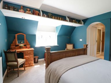 A bedroom with blue walls a wooden bed a wooden dressing table with a chair a window and decorative shelf at Valley House in Devizes