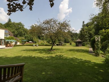 A large lawn with a tree in the center two lounge chairs a wooden bench a house with outdoor seating and umbrellas and two small wooden cottages at Valley House in Devizes