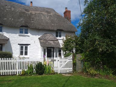 A white cottage with a thatched roof and a white picket fence surrounded by grass and greenery at Furneaux Hatch in Churchstow