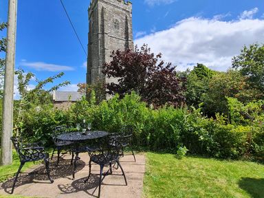 A garden with black metal table and chairs and a stone church tower in the background at Furneaux Hatch in Churchstow