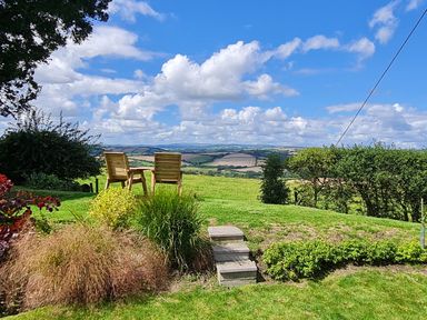 A garden with two wooden chairs and a table overlooking fields and hills at Furneaux Hatch in Churchstow