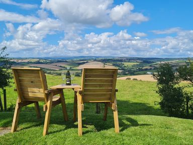 Two wooden chairs with a table holding a bottle and two glasses on grass overlooking rolling fields at Furneaux Hatch in Churchstow