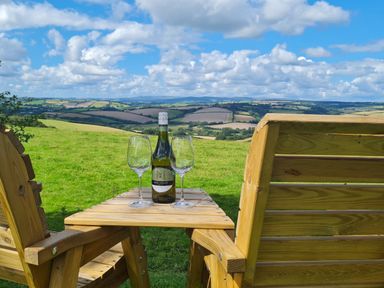 A wooden outdoor table between two chairs with a bottle and two wine glasses overlooking rolling hills at Furneaux Hatch in Churchstow