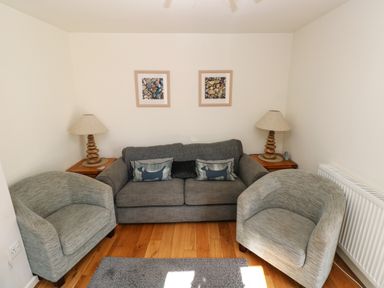 A sitting area with a gray sofa, two gray armchairs, two side tables with lamps, and framed pictures on the wall at Oysterbank Cottage in Llangwm Pembrokeshire