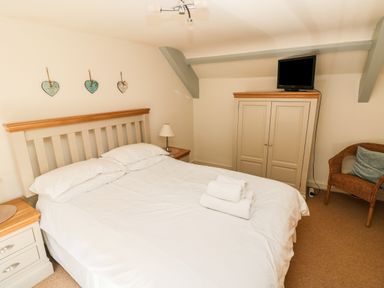 A bedroom with a bed with white linens two bedside tables a wardrobe with a television on top and a wicker chair at Oysterbank Cottage in Llangwm Pembrokeshire