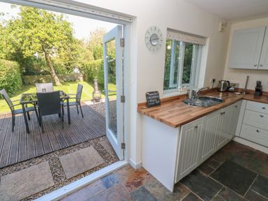 A kitchen with wooden countertops and white cabinets next to an open door leading to a deck with outdoor dining furniture and a garden at Oysterbank Cottage in Llangwm Pembrokeshire