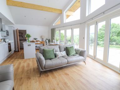 A living room with a gray sofa, green pillows, dining table, kitchen area, and large windows at Pen Bryn in Cyffylliog near Ruthin