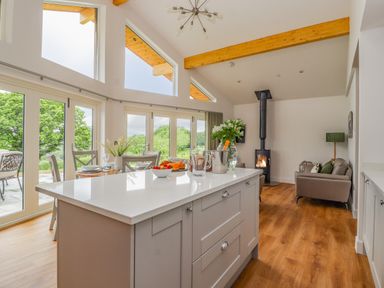 A kitchen island with flowers and fruit with dining table and chairs near glass doors and a living area with armchairs and a wood stove at Pen Bryn in Cyffylliog near Ruthin