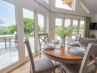 A dining area with a round wooden table set with plates, glasses, and cutlery near large windows overlooking a patio and green landscape at Pen Bryn in Cyffylliog near Ruthin