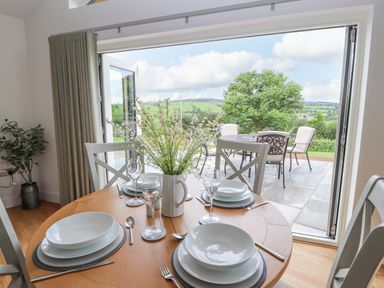 A dining table set with plates glassware and cutlery inside a room with open doors leading to a patio with outdoor chairs and a table and a green landscape at Pen Bryn in Cyffylliog near Ruthin