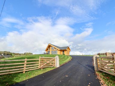 A wooden house at the top of a paved driveway with a wooden gate and green grass at Pen Bryn in Cyffylliog near Ruthin
