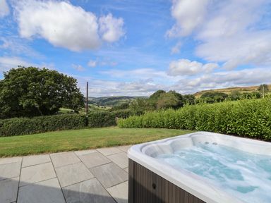 An outdoor hot tub on a stone patio overlooking a lawn and green hedges with hills and trees in the distance at Pen Bryn in Cyffylliog near Ruthin
