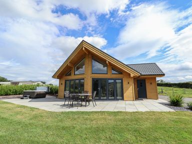 A wooden house with large glass windows patio furniture and a hot tub on a tiled patio with grass in front at Pen Bryn in Cyffylliog near Ruthin