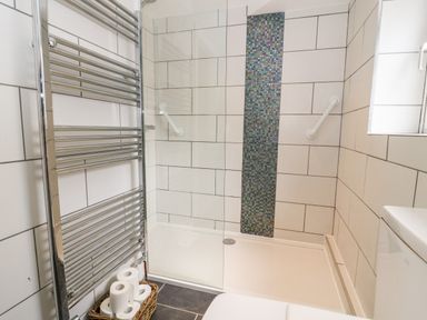 A bathroom with a walk-in shower a tiled wall with a vertical mosaic strip a chrome towel radiator and a basket with toilet paper at Claire Cottage in Gateforth near Hambleton North Yorkshire