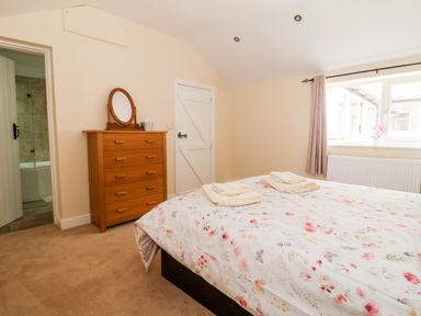 A bedroom with a bed with floral bedding and towels a wooden dresser with a round mirror an open door showing a bathroom and a window with curtains at Claire Cottage in Gateforth near Hambleton North Yorkshire