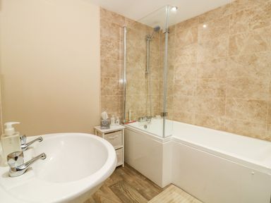 A bathroom with a white sink with soap dispenser a bathtub with glass shower screen and beige tiled walls at Claire Cottage in Gateforth near Hambleton North Yorkshire