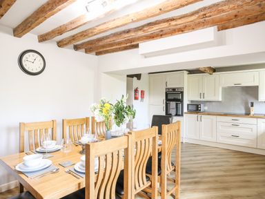 A dining area with a wooden table and chairs set for a meal next to a kitchen with white cabinets and wooden beams on the ceiling at Hafotty Gelynen in Maerdy near Corwen