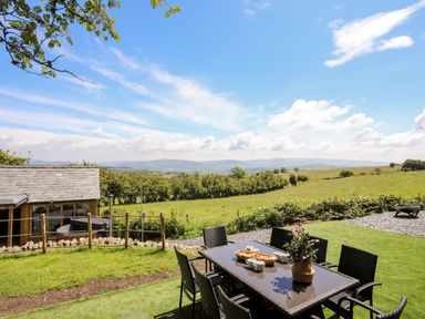 An outdoor patio area with a table, chairs, and a flower pot overlooking fields and hills at Hafotty Gelynen in Maerdy near Corwen
