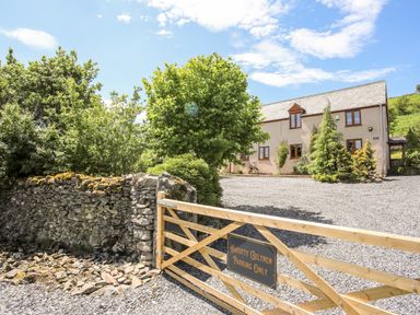 A stone wall and wooden gate with a sign in front of a gravel driveway and a two story house surrounded by trees at Hafotty Gelynen in Maerdy near Corwen