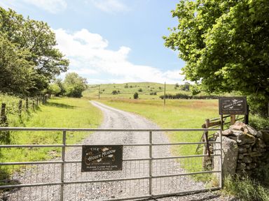 A gravel path behind a metal gate with signs reading The Hidden Meadow at Hafotty Gelynen in a grassy rural area with trees and hills at Hafotty Gelynen Maerdy near Corwen