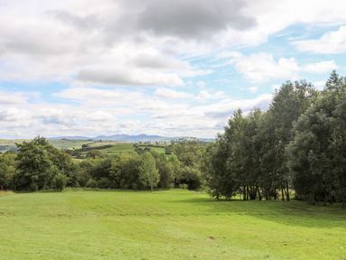 A grassy field with trees on the edges and hills in the background under a partly cloudy sky at Woodland Pod in Maerdy near Corwen