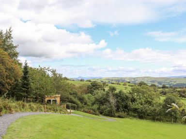A winding gravel path through green grass beside trees and a small wooden structure with hills and cloudy sky in the background at Woodland Pod in Maerdy near Corwen
