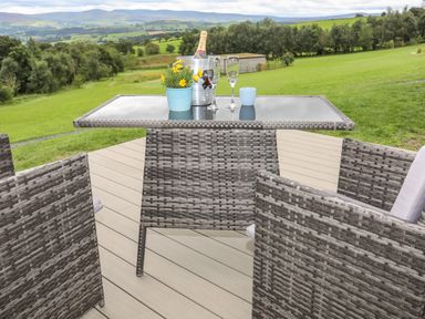 An outdoor patio area with a glass top table and wicker chairs with a champagne bottle in an ice bucket and glasses on the table at Woodland Pod Maerdy near Corwen
