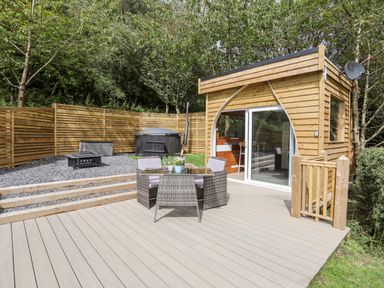 An outdoor deck with a table and chairs next to a wooden cabin and fenced garden with a hot tub and fire pit at Woodland Pod Maerdy near Corwen
