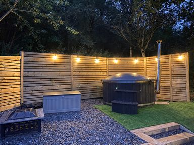 An outdoor area with a hot tub wooden fence string lights and a fire pit at Woodland Pod Maerdy near Corwen