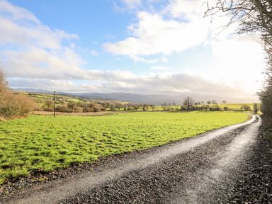 A gravel road curving through green fields with trees and hills under a partly cloudy sky at Woodland Pod in Maerdy near Corwen