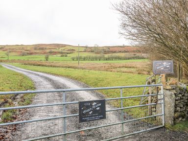 A metal gate with signs for The Hidden Meadow and a gravel driveway leading to green fields and hills at The Hidden Meadow in Maerdy near Corwen